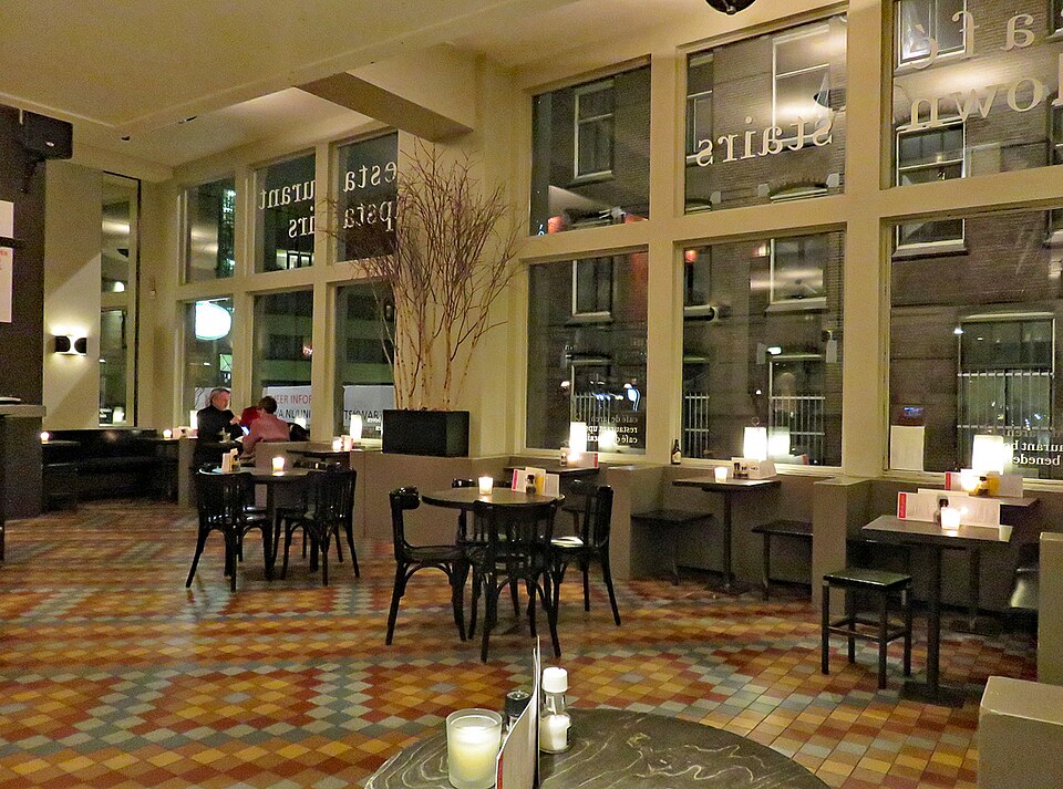 Café tables and chairs on a tiled floor beside tall windows inside Café de Jaren